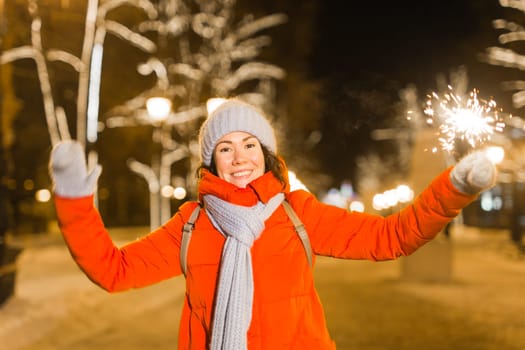 Girl holding a sparkler in her hand. Outdoor winter city background, snow, snowflakes