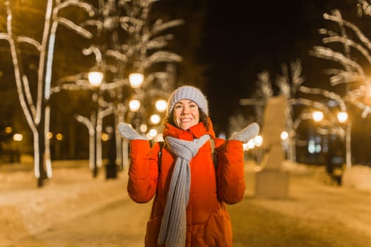 Happy funny young woman with winter clothes background evening city lights illumination. Christmas and winter holidays.
