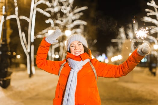 Girl holding a sparkler in her hand. Outdoor winter city background, snow, snowflakes
