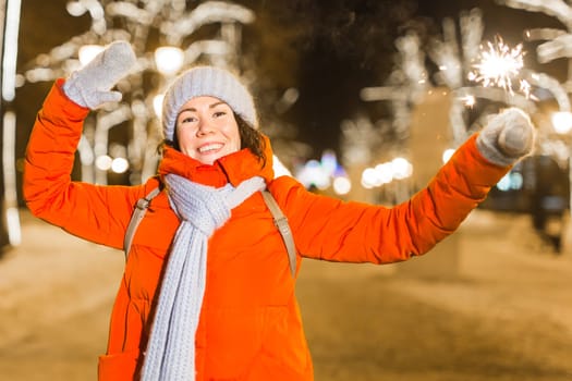 Young beautiful woman in knitted hat and scarf standing in the city with bengal light, sparkler. Concept celebration and christmas