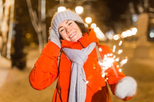 young woman wearing winter knitted clothes holding sparkler outdoors over snow background. Christmas holidays.