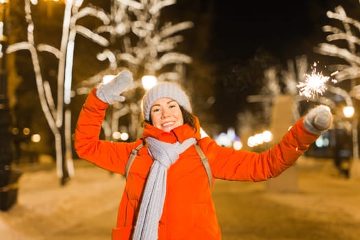 young woman wearing winter knitted clothes holding sparkler outdoors over snow background. Christmas holidays.
