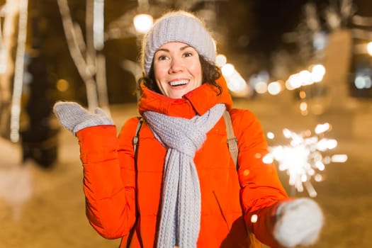 young woman wearing winter knitted clothes holding sparkler outdoors over snow background. Christmas holidays.