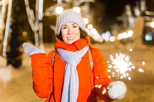 Young beautiful woman in knitted hat and scarf standing in the city with bengal light, sparkler. Concept celebration and christmas