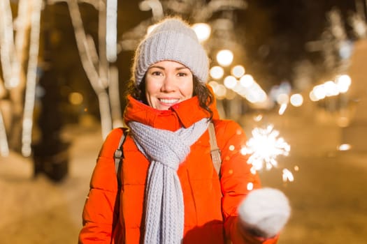 young woman wearing winter knitted clothes holding sparkler outdoors over snow background. Christmas holidays.