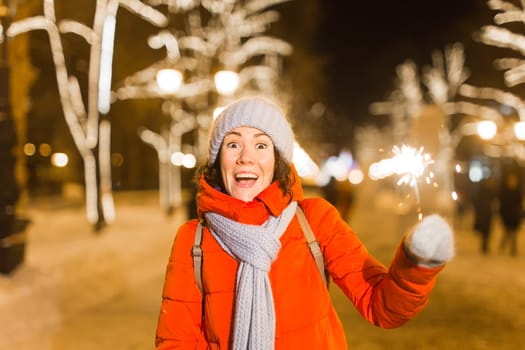 Girl holding a sparkler in her hand. Outdoor winter city background, snow, snowflakes