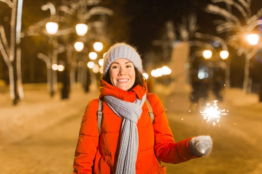 young woman wearing winter knitted clothes holding sparkler outdoors over snow background. Christmas holidays.