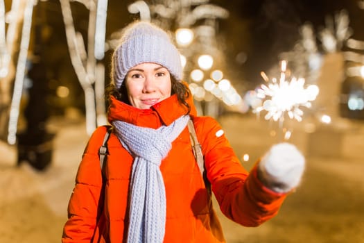 young woman wearing winter knitted clothes holding sparkler outdoors over snow background. Christmas holidays.
