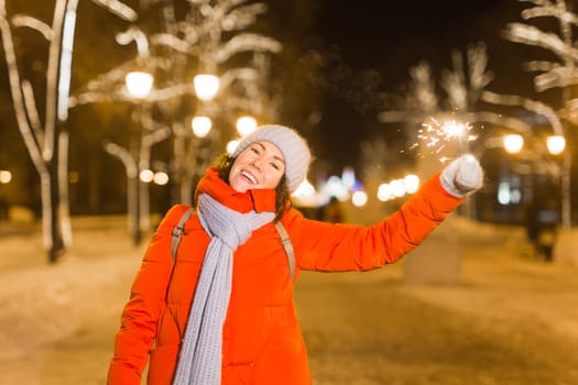 Young beautiful woman in knitted hat and scarf standing in the city with bengal light, sparkler. Concept celebration and christmas
