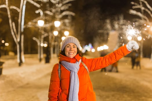 young woman wearing winter knitted clothes holding sparkler outdoors over snow background. Christmas holidays.