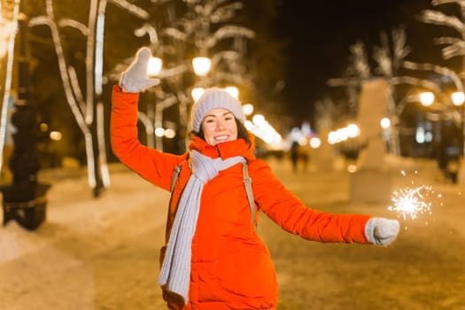 Young beautiful woman in knitted hat and scarf standing in the city with bengal light, sparkler. Concept celebration and christmas