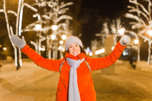 Girl holding a sparkler in her hand. Outdoor winter city background, snow, snowflakes