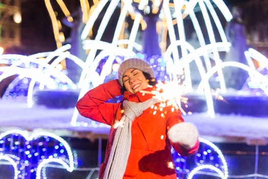 Young beautiful woman in knitted hat and scarf standing in the city with bengal light, sparkler. Concept celebration and christmas