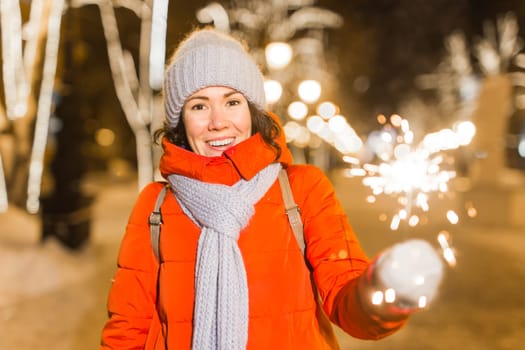 young woman wearing winter knitted clothes holding sparkler outdoors over snow background. Christmas holidays.