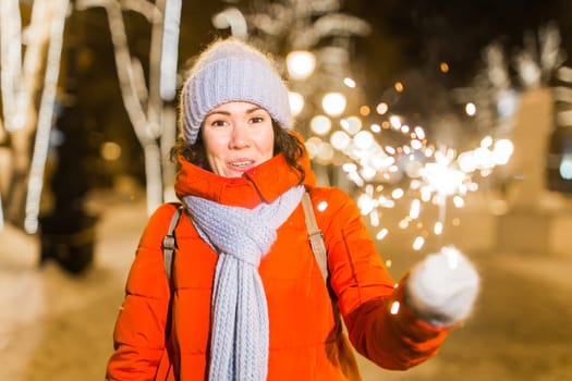 Young beautiful woman in knitted hat and scarf standing in the city with bengal light, sparkler. Concept celebration and christmas