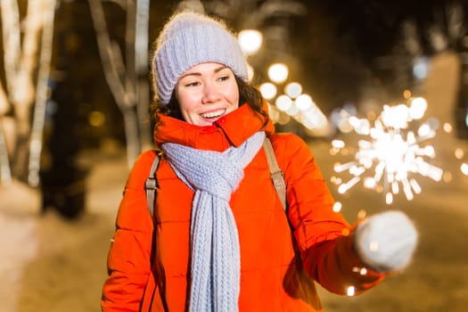 Young beautiful woman in knitted hat and scarf standing in the city with bengal light, sparkler. Concept celebration and christmas