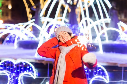 Girl holding a sparkler in her hand. Outdoor winter city background, snow, snowflakes