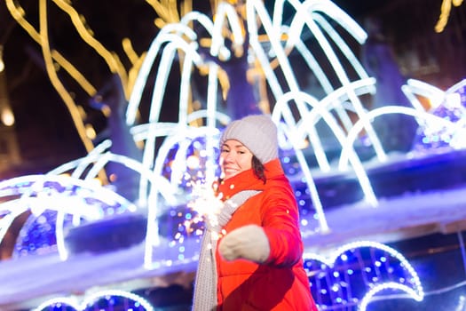 young woman wearing winter knitted clothes holding sparkler outdoors over snow background. Christmas holidays.