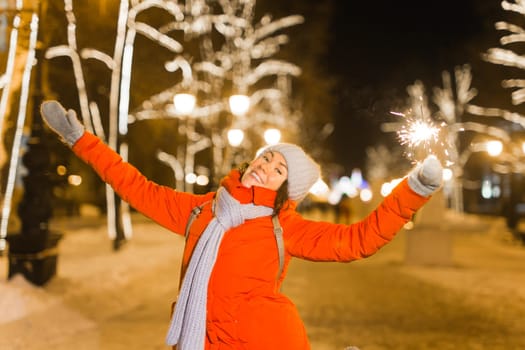 young woman wearing winter knitted clothes holding sparkler outdoors over snow background. Christmas holidays.