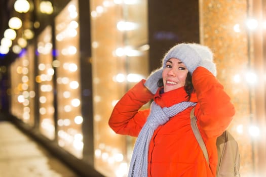 Girl having fun christmas decoration lights street. Young happy smiling woman wearing stylish knitted scarf and jacket outdoors. Model laughing. Winter wonderland city scene, New Year party.