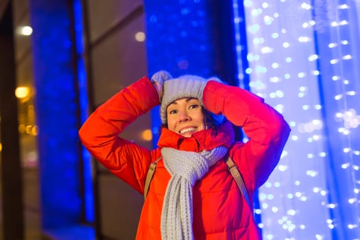 Girl having fun christmas decoration lights street. Young happy smiling woman wearing stylish knitted scarf and jacket outdoors. Model laughing. Winter wonderland city scene, New Year party.