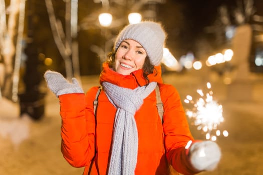 young woman wearing winter knitted clothes holding sparkler outdoors over snow background. Christmas holidays.