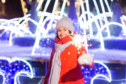 Young beautiful woman in knitted hat and scarf standing in the city with bengal light, sparkler. Concept celebration and christmas