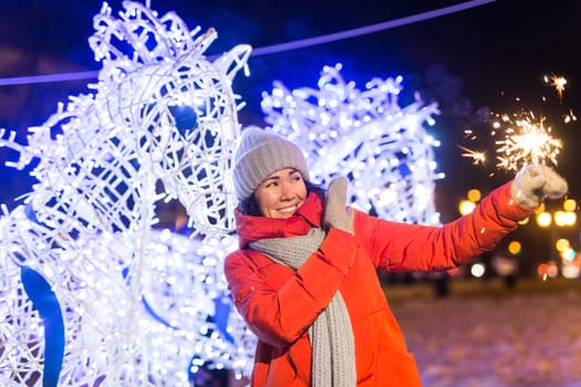 Young beautiful woman in knitted hat and scarf standing in the city with bengal light, sparkler. Concept celebration and christmas