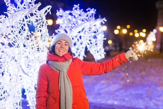 young woman wearing winter knitted clothes holding sparkler outdoors over snow background. Christmas holidays.
