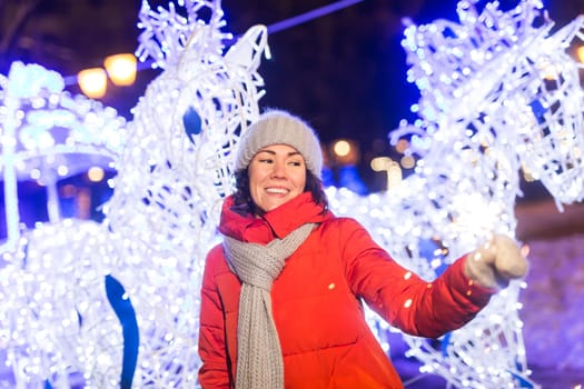 young woman wearing winter knitted clothes holding sparkler outdoors over snow background. Christmas holidays.