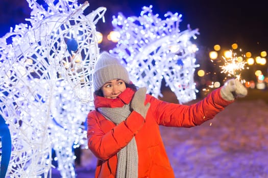 young woman wearing winter knitted clothes holding sparkler outdoors over snow background. Christmas holidays.