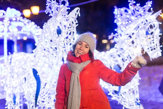 young woman wearing winter knitted clothes holding sparkler outdoors over snow background. Christmas holidays.