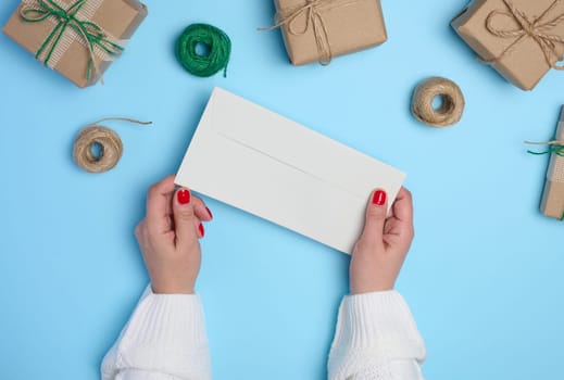 Female hands holding a white paper envelope on a blue background, around a box with gifts, top view