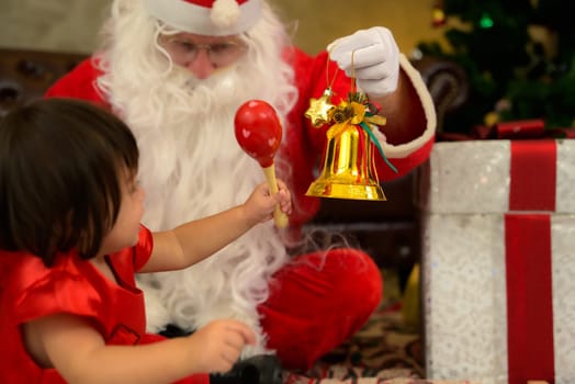 Little kid girl siiting together with Santa claus on floor and playing with toy bell at home. Celebrate holiday Christmas and Thanksgiving party.