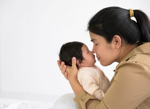 Asian mother holding newborn girl in her hands and gently kissing baby nose with love. Happy mom sitting on white bed at home. Safe with mom hug and healthy newborn baby concept