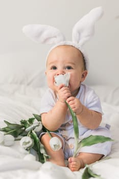 Cute funny baby with bunny ears, with a tulips at home on a bed of white bedding. Easter greeting card.