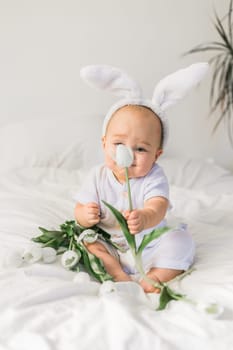 Cute funny baby with bunny ears, with a tulips at home on a bed of white bedding. Easter greeting card.