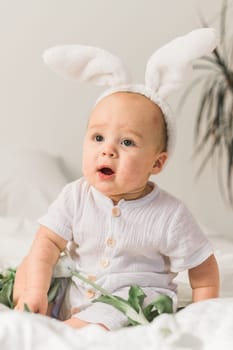 Cute funny baby with bunny ears, with a tulips at home on a bed of white bedding. Easter greeting card.