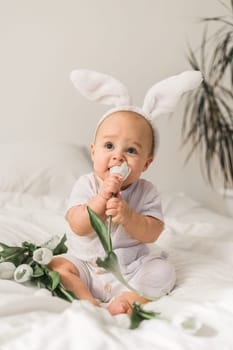 Cute funny baby with bunny ears, with a tulips at home on a bed of white bedding. Easter greeting card.
