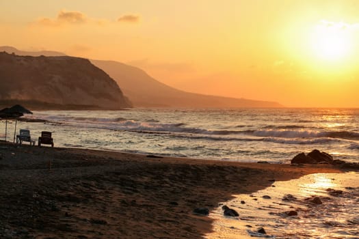 Sunset shines to two sunbeds on empty wild beach. Small sea waves, hills in hazy background