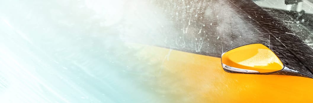 Side mirror and window of orange vehicle washed in self serve car wash, left part filled with abstract shampoo streaks on glass - wide carwash banner, copy space for text.