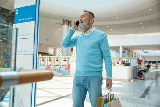 Stylish senior man with phone walking with purchases. Consumerism, shopping and retired people lifestyle concept