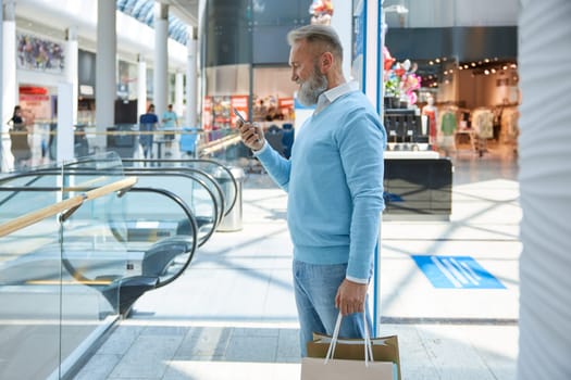 Rich senior old man booking taxi online standing with shopping bags in supermarket. Elderly grandfather tourist in city mall