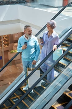 Senior couple with purchases in bags riding down shopping mall escalator enjoying time together