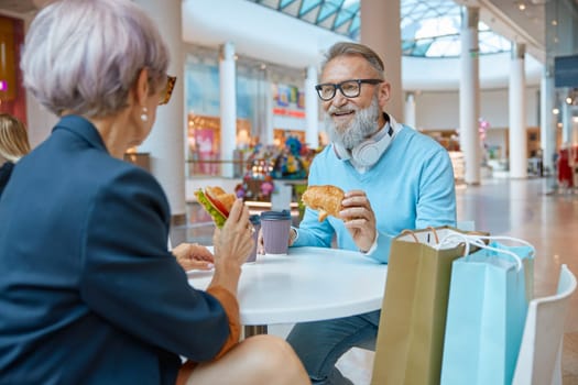 Happy retired couple in love eating croissants at cafe in shopping mall. Successful senior people and leisure activity on weekends concept