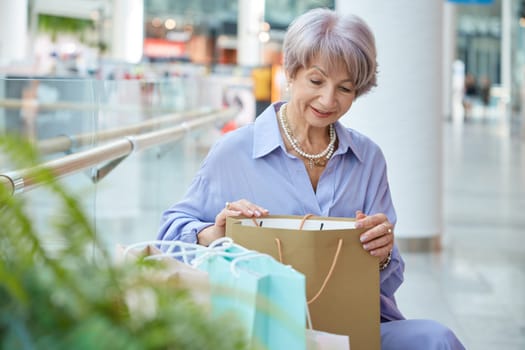 Fashion elderly woman with purchases sitting on bench in shopping mall feeling surprised to find little gift from favorite brand