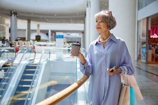 Senior woman with cup of coffee and shopping bags over mall store showcase. Stylish grandmother feeling satisfied enjoying discounts in shop