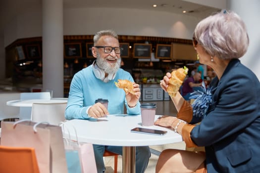 Overjoyed retired couple in love eating croissant at cafeteria in shopping mall. Successful senior people and leisure activity on weekends concept