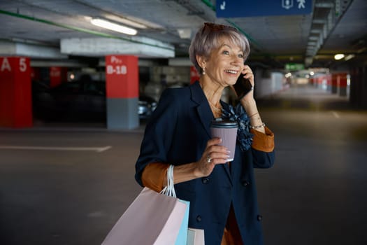 Happy elderly woman talking mobile phone walking through parking of shopping mall. Senior lady sharing expression after successful purchases with low prices