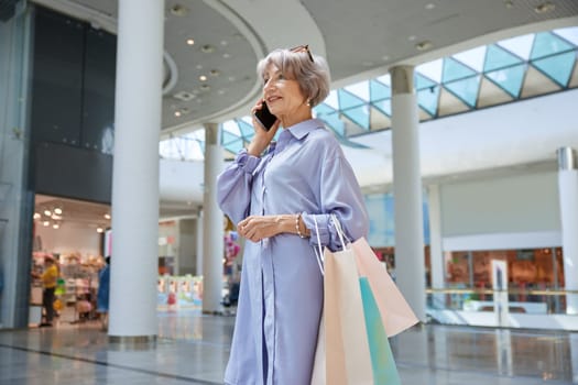 Portrait of attractive senior woman with phone and shopping bags over mall store showcase. Stylish grandmother feeling satisfied enjoying discounts in shop